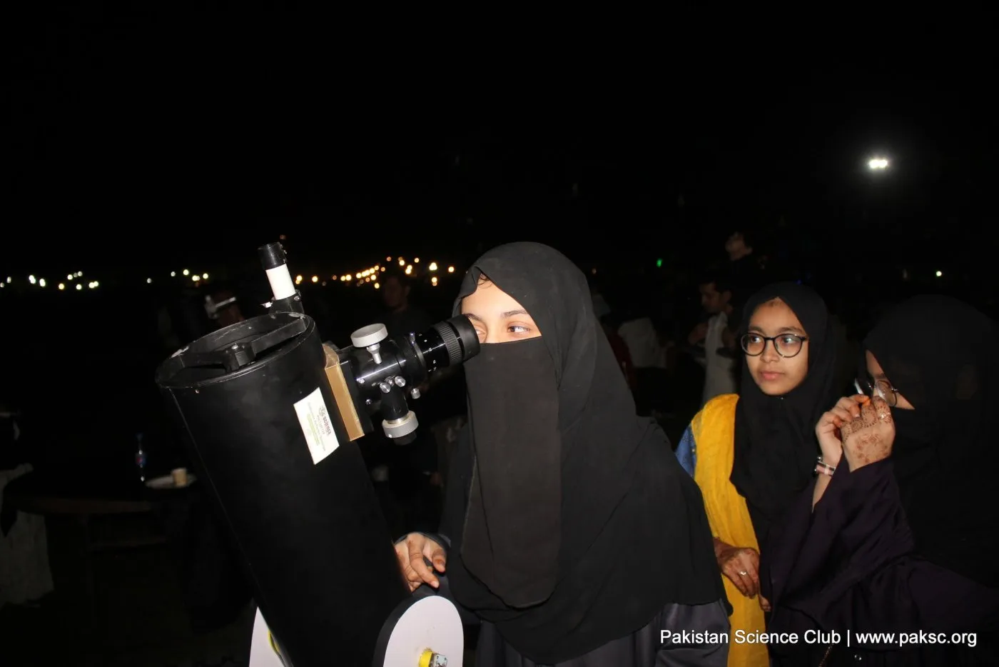 A visitor looks through the eyepiece of a Dobsonian telescope at night while others wait beside her.