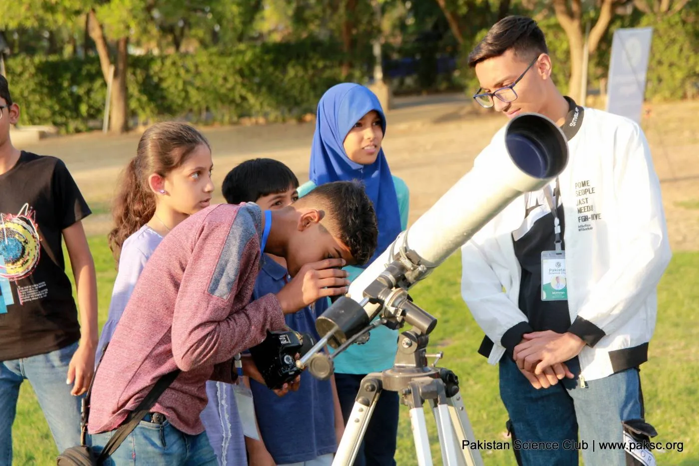 A boy looks through a white refractor telescope on a tripod while other children and a volunteer wait their turn.