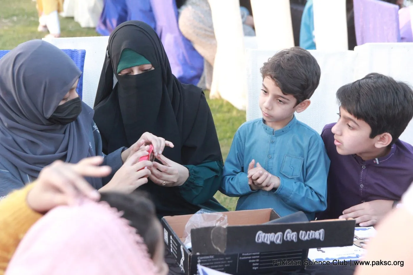 Two boys and two women assemble small telescope kits from a box at a Pakistan Science Club telescope-making workshop on a lawn.