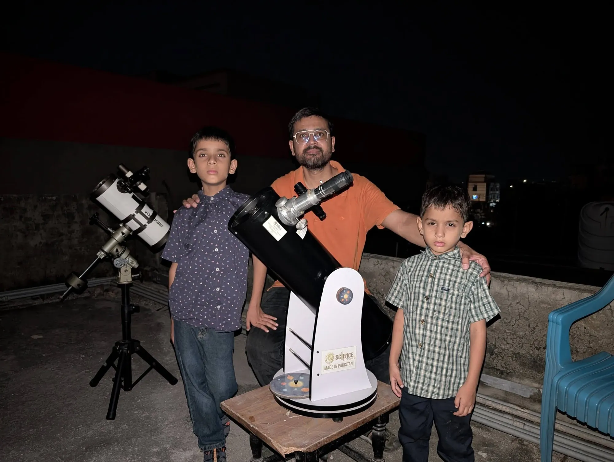 A man and two young boys on a rooftop at night beside a Made in Pakistan Dobsonian telescope, with a white reflector telescope on a tripod next to them.